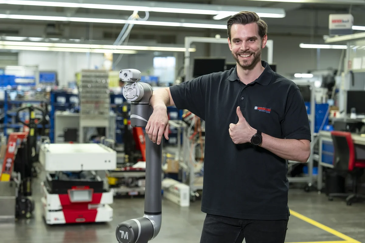 Gessmannn Mitarbeiter lacht, stützt sich auf Cobot und zeigt Daumen nach oben.